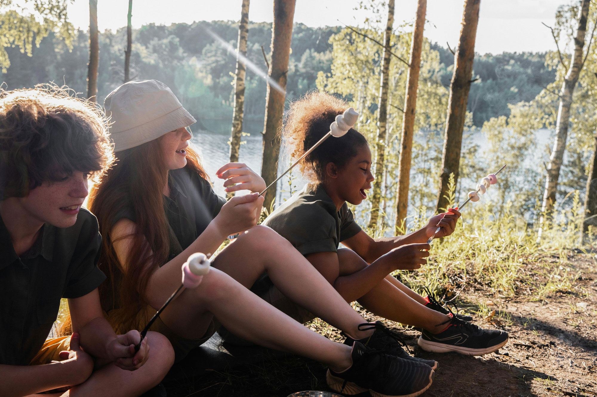 Three kids sit in a sunlit forest, smiling and roasting marshmallows on sticks.
